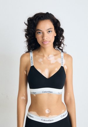 Zwarte bralette met een v-hals ontwerp, voorzien van dunne bandjes en een witte "Calvin Klein" logo band aan de onderkant. Gladde textuur, minimalistische stijl.