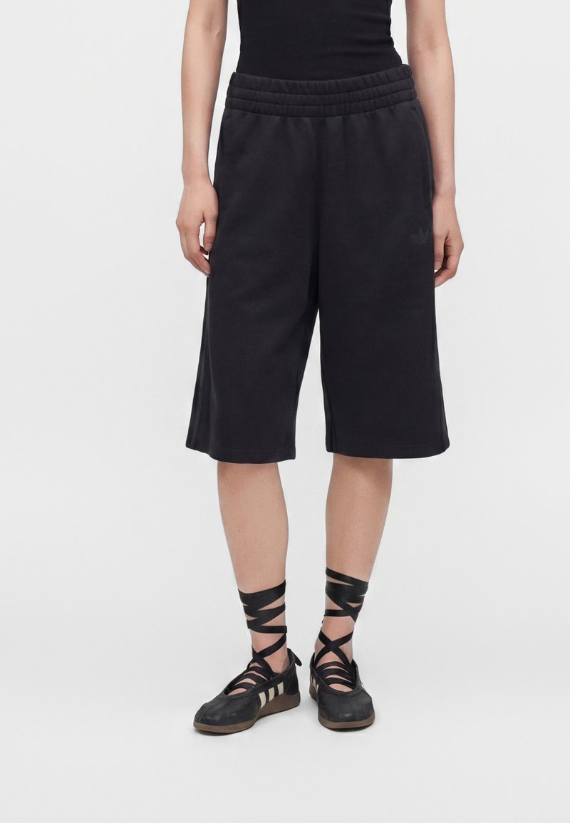 Personne portant un short noir large arrivant au genou et des chaussures de style ballerine noires avec des rubans croisés à la cheville.