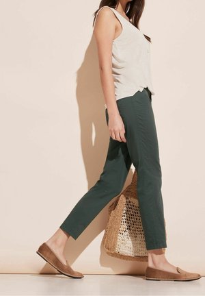 Nainen kävelee pukeutuneena hihattomaan beigeen toppiin, tummanvihreisiin lyhytlahkeisiin housuihin, ruskeisiin slip-on kenkiin, kantaa kudottua kantolaukkua beigeä taustaa vasten.