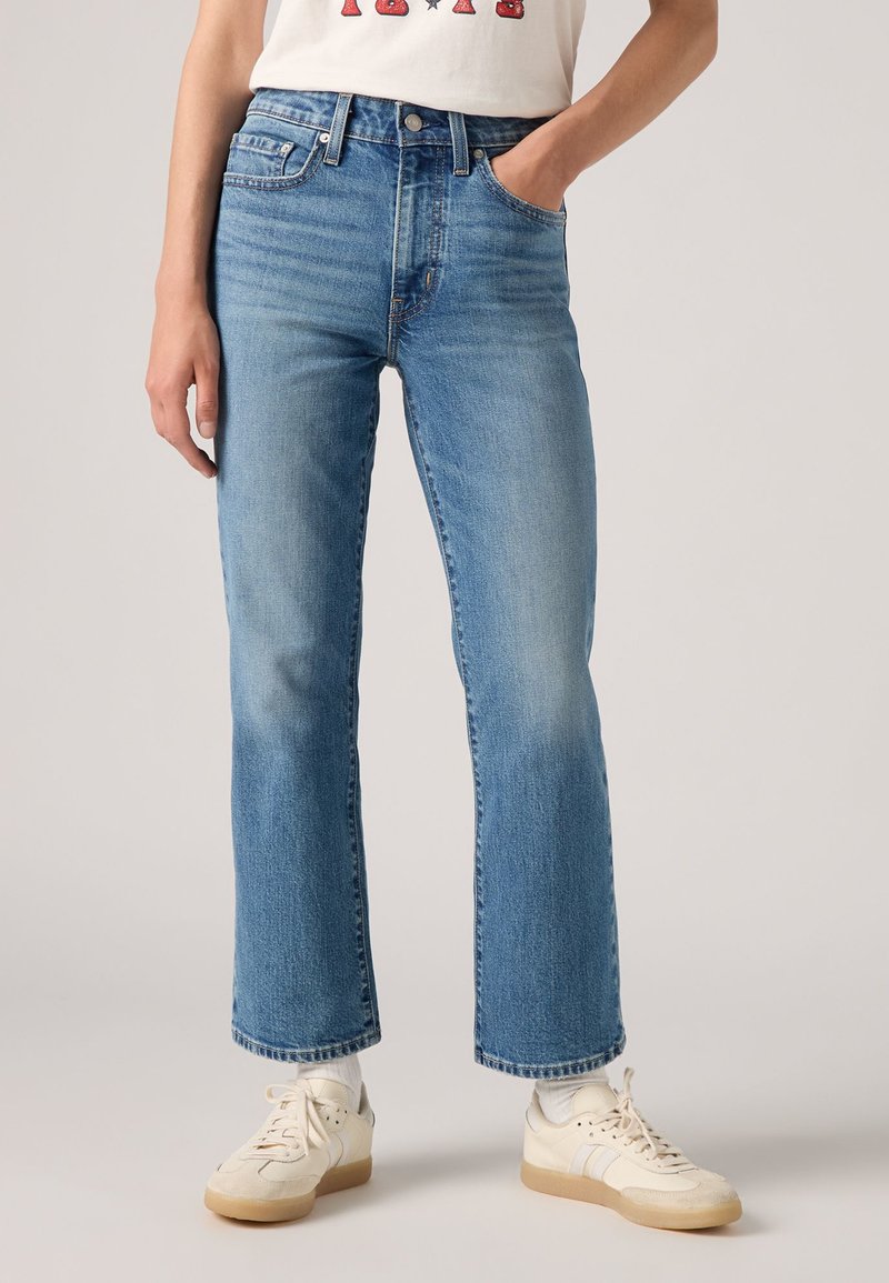 Jean en denim bleu clair avec une coupe droite, taille haute, design à cinq poches et ourlet effiloché. Portés avec des baskets blanches.