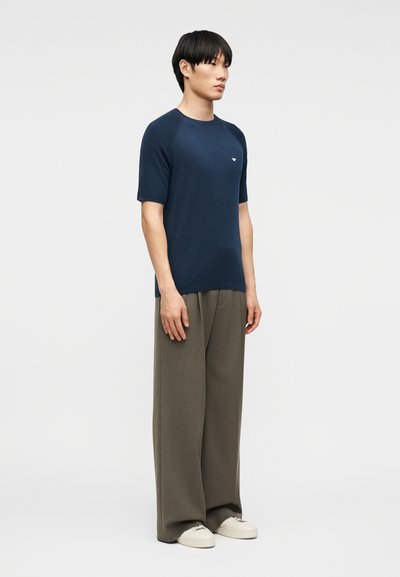 Νεαρός άνδρας που στέκεται, φοράει μπλε κοντομάνικο πουκάμισο, φαρδιά χακί παντελόνια και λευκά αθλητικά παπούτσια, μπροστά σε απλό φωτεινό φόντο.