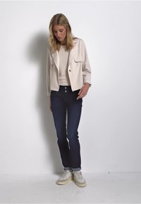 Beige bunda se dvěma předními kapsami přes světlý top, spárovaná s tmavými džíny a béžovými teniskami. Jednoduchý design, hladká textura, bez vzorů.