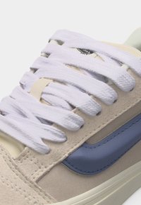 Κοντινό πλάνο μπεζ αθλητικού παπουτσιού με φαρδιά λευκά κορδόνια και μπλε λωρίδα στο πλάι, σε λευκό φόντο.