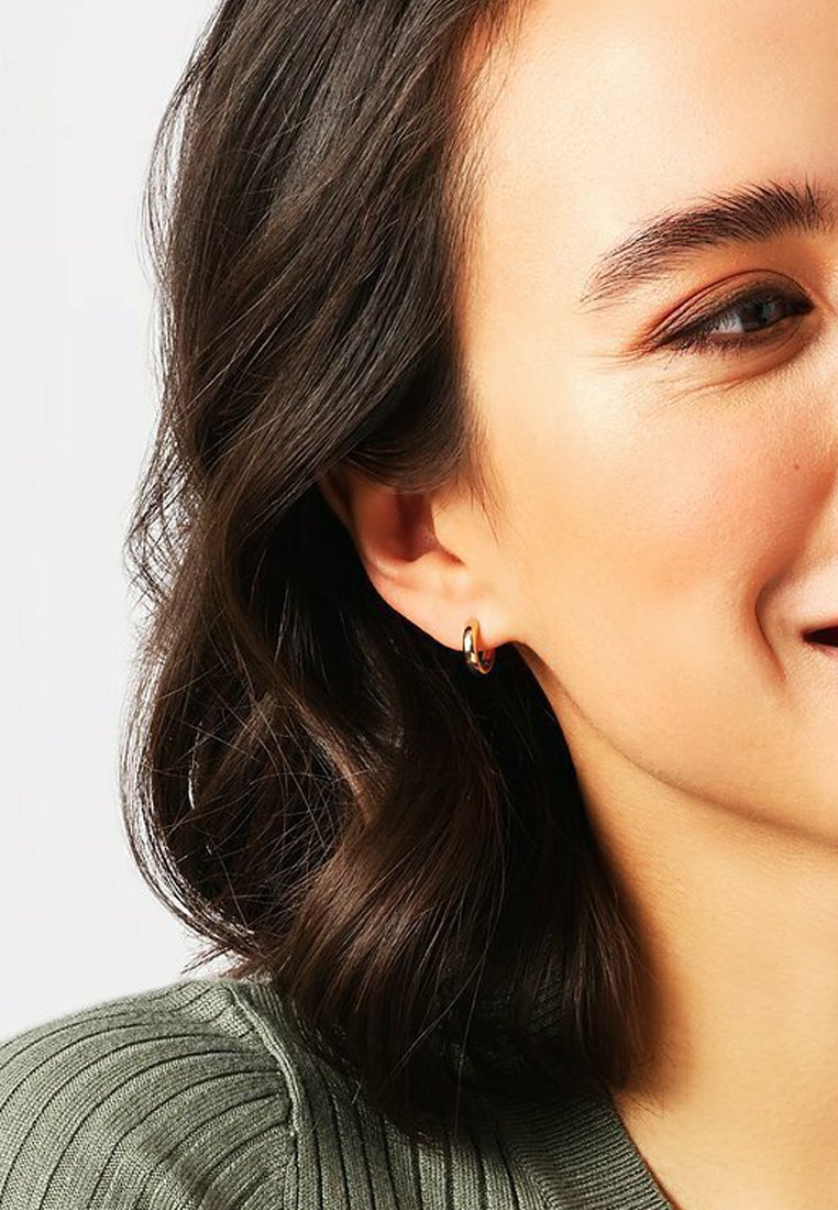 Gli orecchini a cerchio in oro presentano una finitura liscia, forma circolare e un design sottile. Indossati da un modello con capelli scuri e ondulati e un maglione verde lavorato a maglia.