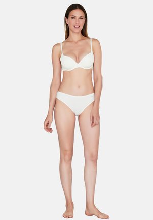 Kvinde står barfodet og vender fremad, iført en hvid bh og matchende hvide underbukser mod en ensfarvet baggrund.