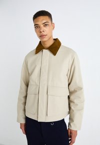 Chaqueta de algodón beige con un cuello de pana marrón, dos bolsillos frontales y cierre con cremallera, con un corte recto y un diseño minimalista.