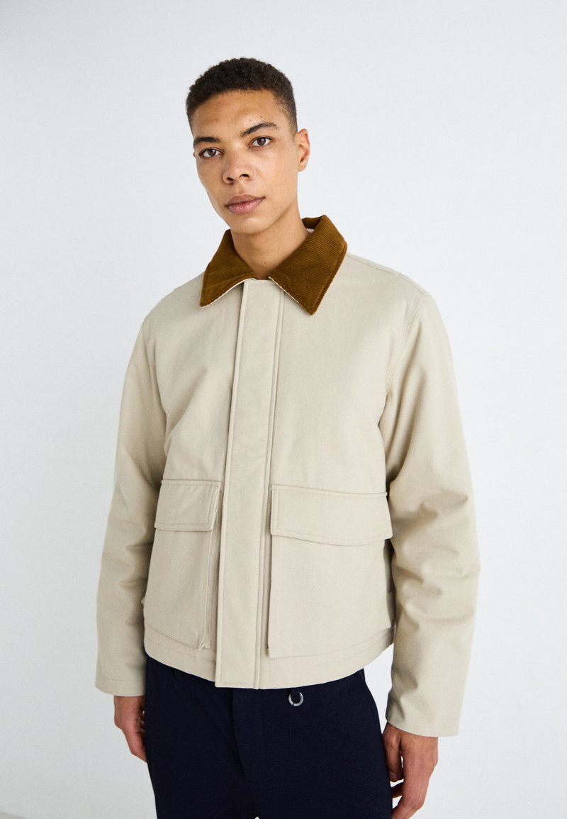 Chaqueta de algodón beige con un cuello de pana marrón, dos bolsillos frontales y cierre con cremallera, con un corte recto y un diseño minimalista.