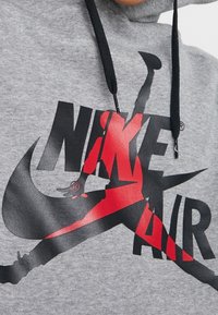 Sudadera gris de felpa con un logo de Nike Air en blanco y rojo. Presenta una silueta gráfica y cordones negros en la capucha.