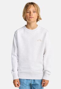 Felpa bianca con colletto rotondo, polsini e orlo a costine. Presenta un piccolo logo ricamato in beige. Indossata con jeans azzurri chiari.