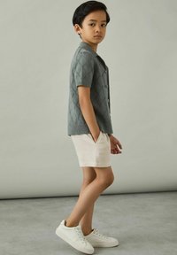 Grå stickad kortärmad cardigan med diamantmönster och knappar, i kombination med beige shorts och vita sneakers, visad stående på sidan.