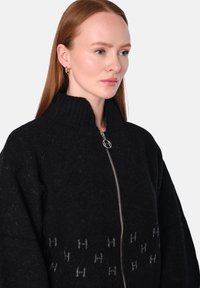 Svart ull zip-up tröja med hög ribbad krage, med ett subtilt grått mönster av 'H'-bokstäver över framsidan. Mjuk textur, oversized passform.