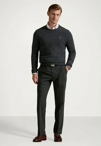 Man står klädd i en mörkgrå tröja över en vit skjorta med krage, grå byxor, bruna skor och en armbandsklocka, poserar med en hand i fickan.