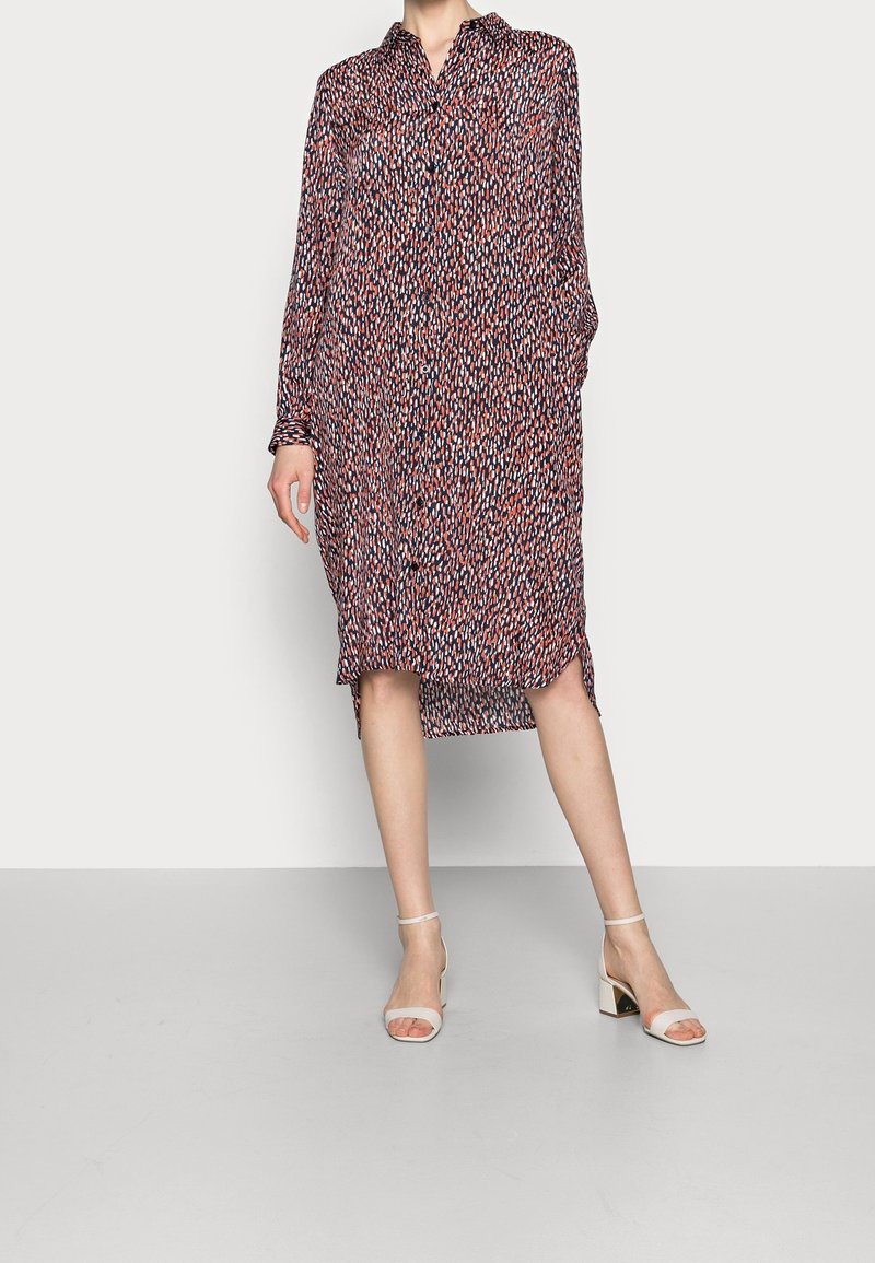 Femme portant une robe midi ample à manches longues boutonnée avec un motif abstrait multicolore et des talons blocs blancs ouverts à l'avant.