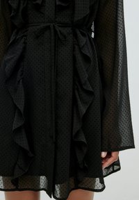 Schwarzes Kleid aus leichtem Stoff mit Rüschendetails, einem Taillengürtel und gepunkteter Textur; durchsichtige Ärmel und Saum.