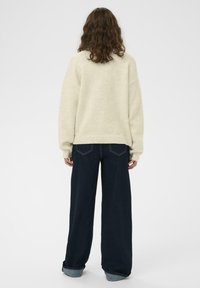 Off-white oversized pulover s teksturiranim završetkom, uparen s širokim tamnim trapericama, koje imaju preokrenute manžete i detalje na stražnjim džepovima.