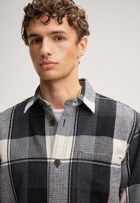 Jonge man met krullend haar, gekleed in een zwart-wit geblokt overhemd over een wit T-shirt, neutrale achtergrond.