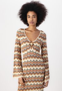 Robe en tricot à manches longues avec un motif à chevrons beige, brun, orange et gris, dotée d'un décolleté en V et d'un détail en métal circulaire au niveau de la poitrine.