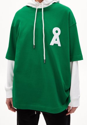 Osoba nosząca na sobie warstwową, zieloną, oversize'ową koszulkę na białej bluzie z kapturem, na piersi znajduje się biały logotyp Å, a sznurki kaptura są białe.