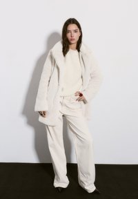 Fehér műszőrme kabát széles hajtókákkal, krémszínű pulóverrel és világos krémszínű, széles szárú nadrággal párosítva. Fekete, hegyes orrú magassarkú cipők. Egyszerű ékszerek.