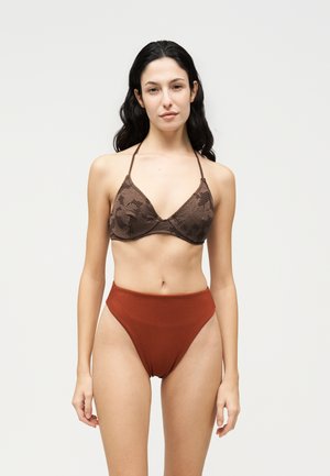 Une femme porte un soutien-gorge marron en dentelle à tour de cou et une culotte taille haute couleur rouille, sur un fond clair et uni.