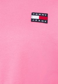 Tessuto di cotone rosa con superficie texturizzata, presenta un'etichetta rettangolare con il testo "TOMMY JEANS" e un logo in rosso, bianco e blu.