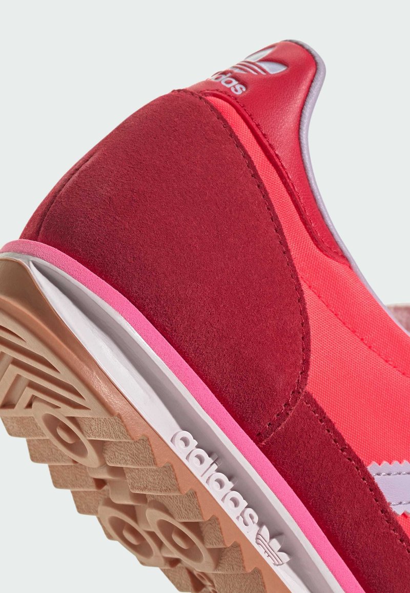 Zapatilla deportiva roja y rosa con parte superior de ante, paneles sintéticos en contraste, logo blanco en el talón y suela de goma texturizada.
