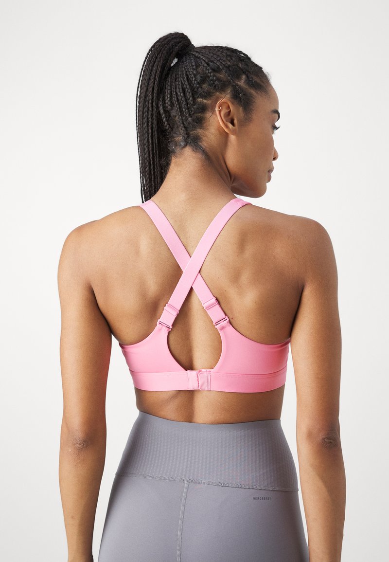 Rosa Sport-BH mit überkreuzten Trägern, der eine glatte Textur und einen verstellbaren Verschluss aufweist. Graue hochtaillierte Leggings, die am Bund teilweise sichtbar sind.