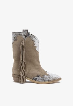 Bota estilo occidental en ante beige con una puntera decorativa plateada y detalle de flecos en la parte trasera. Presenta acentos metálicos y costuras.