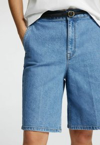 Jeansshorts i medelblått, med en avslappnad passform, två sidofickor och ett svart bälte med ett gyllene spänne. Avslutningen inkluderar kontrastsöm.
