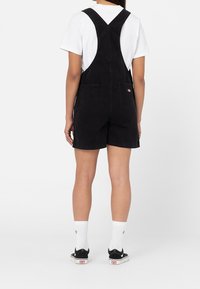 Černé denimové šortkové overaly s nastavovacími popruhy, dvěma zadními kapsami a designem šortek. Oblečeny s bílým tričkem a černými teniskami.