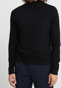 Pull à col montant noir en maille fine, avec des manches longues et une coupe ajustée, associé à un pantalon bleu marine.