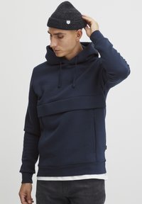 Felpa con cappuccio di un blu navy scuro, caratterizzata da un design a strati, tessuto testurizzato e una tasca frontale. Accoppiata con un berretto scuro.