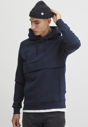 Hoodie u tamnoplavoj boji, s višeslojnim dizajnom, teksturiranim materijalom i prednjim džepom. Uparen s tamnom kapom.