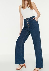 Jeans in denim scuro a vita alta e gamba ampia, con cinque bottoni in rame, tasche laterali e una texture liscia.