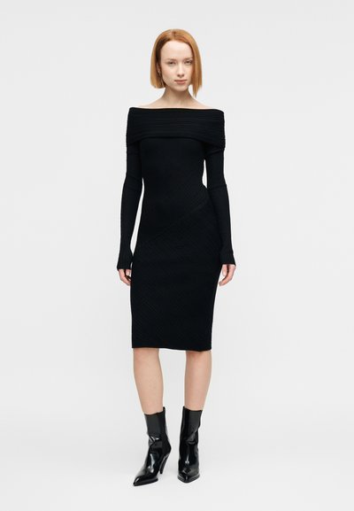 Γυναίκα με κοντά κόκκινα μαλλιά, φορώντας στενή μαύρη off-shoulder φόρεμα και μαύρα μποτάκια με τακούνι, στέκεται μπροστά σε ένα λευκό φόντο.