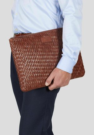 Mann i blå skjorte og mørke bukser som holder en stor brun flettet lærveske med glidelås og synlig merkeetikett.