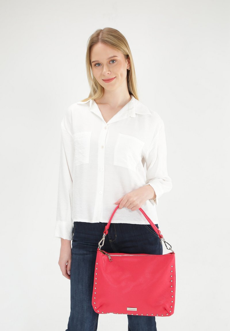 Rote Lederhandtasche mit silbernen Nieten, Reißverschluss und einem einzigen Henkel, gehalten von einer Person in einem weißen Hemd und dunklen Jeans vor einem neutralen Hintergrund.