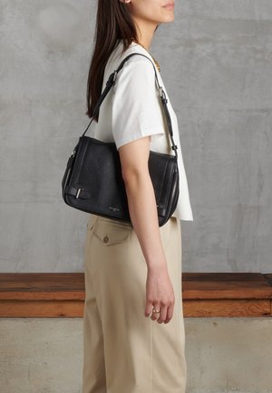 Borsa a tracolla nera in pelle con finitura testurizzata, cerniere laterali e una tracolla removibile, indossata da una persona con una camicia chiara.