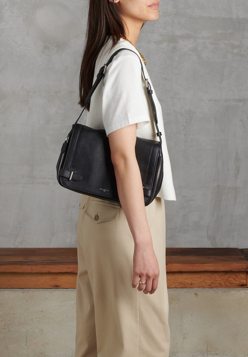 Schwarze Leder-Crossbody-Tasche mit strukturiertem Finish, seitlichen Reißverschlüssen und abnehmbarem Schultergurt, getragen von einer Person mit einem hellen Hemd.
