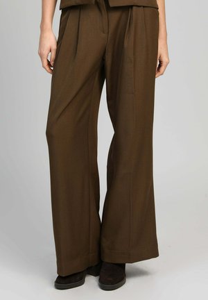 Personne portant un pantalon large marron et des chaussures marron foncé, debout devant un fond gris uni.