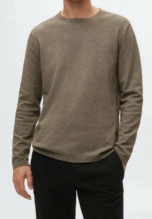 Mies, joka pukeutunut yksiväriseen oliivinvihreään pitkähihainen pyöreäkauluspuseroon ja mustiin housuihin, seisoo käsi yhdessä taskussa.