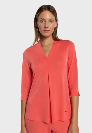 Korallinvärinen pusero, jossa on kolme neljäsosan hihaa, V-kaula-aukko ja laskosyksityiskohta. Valmistettu sileästä kankaasta, jossa on hienoinen kiilto ja hillitty brändäys.