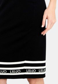 Gonna nera a maglia con strisce orizzontali bianche e logo "LIU JO". Primo piano dell'orlo con una mano posizionata vicino al tessuto.