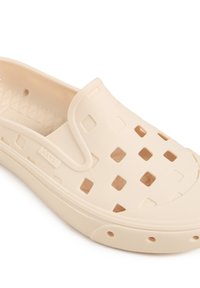 Chaussures beiges claires à enfiler, en caoutchouc flexible, avec un design perforé présentant des découpes carrées et des semelles texturées pour l'adhérence.