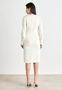 Beiges, tailliertes Kleid mit langen Ärmeln, Reißverschluss hinten und maßgeschneiderten Nähten. Glattes Material und knielanger Schnitt. Neutrale Farbe ergänzt verschiedene Stile.