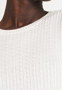 Vit stickad tröja med en texturerad ribbad design och rundad halsringning, som framhäver mjukt, lätt tyg med en subtil glans.