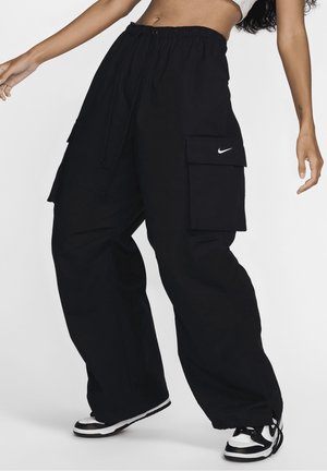 Kobieta ubrana w luźne czarne spodnie cargo z kieszeniami i ściągaczem w talii, w zestawie z czarno-białymi sneakersami, stojąca na białym tle.