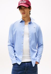 Ung mann iført rød cap, lyseblå skjorte over en hvit singlet, og mørkeblå jeans med hendene i lommene.