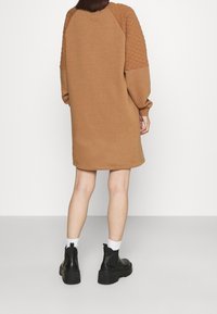 Hnědá sweatshirt šaty s prošívanými rameny, dlouhými rukávy a volným střihem, doplněné černými kotníkovými botami a bílými ponožkami.