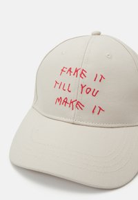 Cappellino da baseball beige con testo ricamato rosso "FAKE IT TILL YOU MAKE IT"; design strutturato, visiera curva e dettagli cuciti.